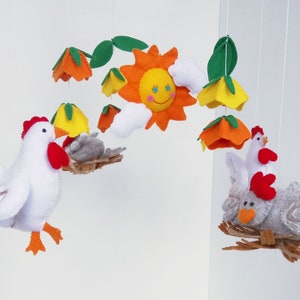 K&ouml;nnte beinhalten: Ein Filz-Mobile mit einer gelben Sonne, wei&szlig;en Wolken, orangefarbenen und gelben Blumen und zwei wei&szlig;en H&uuml;hnern mit roten K&auml;mmen. Ein Huhn sitzt in einem Nest mit einem grauen K&uuml;ken.