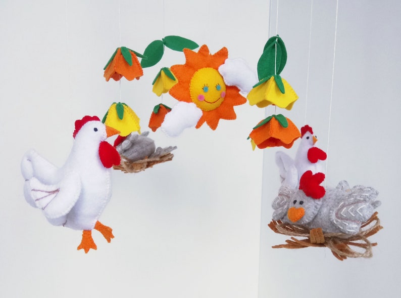 K&ouml;nnte beinhalten: Ein Filz-Mobile mit einer gelben Sonne, wei&szlig;en Wolken, orangefarbenen und gelben Blumen und zwei wei&szlig;en H&uuml;hnern mit roten K&auml;mmen. Ein Huhn sitzt in einem Nest mit einem grauen K&uuml;ken.