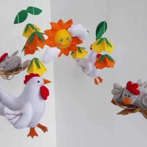 K&ouml;nnte beinhalten: Ein Mobile aus Filz mit einem wei&szlig;en Huhn mit orangenen F&uuml;&szlig;en und einem roten Kamm, einer gelben Sonne, gr&uuml;nen Bl&auml;ttern und zwei grauen Filzv&ouml;geln.