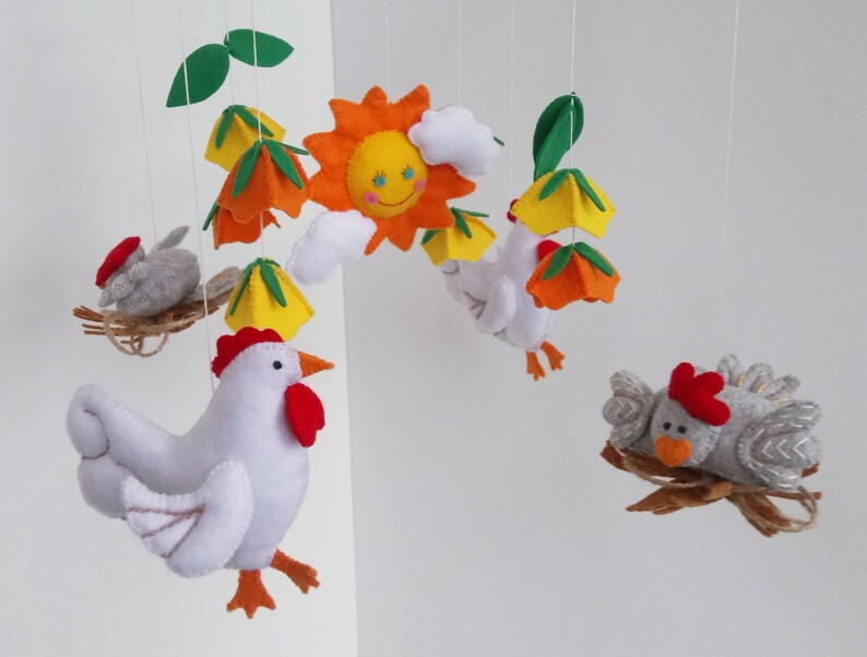 K&ouml;nnte beinhalten: Ein Mobile aus Filz mit einem wei&szlig;en Huhn mit orangenen F&uuml;&szlig;en und einem roten Kamm, einer gelben Sonne, gr&uuml;nen Bl&auml;ttern und zwei grauen Filzv&ouml;geln.