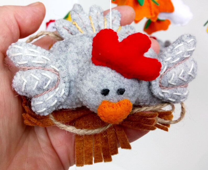 K&ouml;nnte beinhalten: Ein graues Filzh&auml;hnchen mit rotem Filzkamm und orangefarbenem Filzschnabel, das in einem braunen Filznest mit ausgefransten R&auml;ndern sitzt.