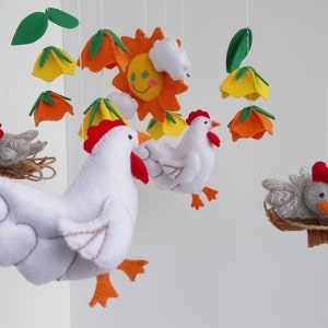 K&ouml;nnte beinhalten: Ein Mobile aus wei&szlig;em Filz mit einem Huhn, einer gelben, orangen und gr&uuml;nen Filzblume, einem grauen Filz-K&uuml;ken in einem Nest und einer gelben Filzsonne mit einem l&auml;chelnden Gesicht.