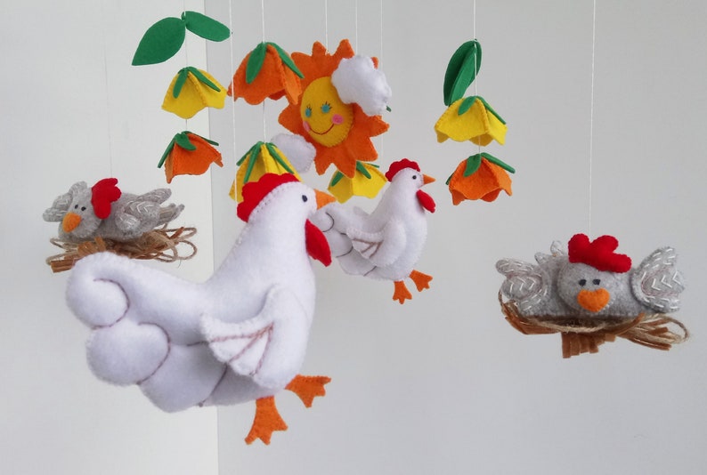 K&ouml;nnte beinhalten: Ein Mobile aus wei&szlig;em Filz mit einem Huhn, einer gelben, orangen und gr&uuml;nen Filzblume, einem grauen Filz-K&uuml;ken in einem Nest und einer gelben Filzsonne mit einem l&auml;chelnden Gesicht.