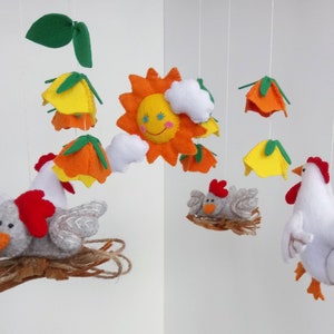 K&ouml;nnte beinhalten: Ein Filz-Babymobil mit einer gelben Sonne, orangefarbenen und gelben Blumen, wei&szlig;en Wolken und zwei grauen K&uuml;ken in Nestern. Ein wei&szlig;es Huhn mit rotem Kamm h&auml;ngt darunter.