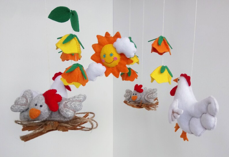 K&ouml;nnte beinhalten: Ein Filz-Babymobil mit einer gelben Sonne, orangefarbenen und gelben Blumen, wei&szlig;en Wolken und zwei grauen K&uuml;ken in Nestern. Ein wei&szlig;es Huhn mit rotem Kamm h&auml;ngt darunter.