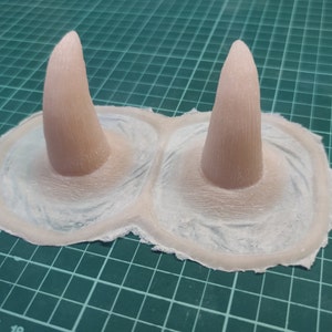 Puede incluir: Dos cuernos de silicona de color rosa pálido con una superficie texturizada, colocados sobre una estera de corte verde.