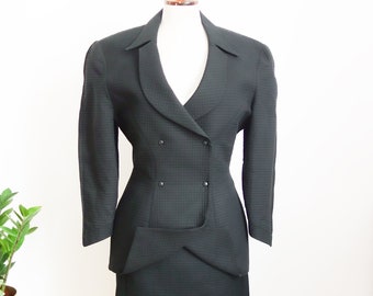 May include: Black two-piece suit with a double-breasted blazer and a matching skirt. The suit has a textured pattern and is displayed on a mannequin. The blazer features a notched lapel and two rows of buttons.