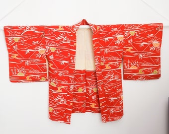 May include: A red kimono with a white and gold landscape pattern. The kimono is hanging on a white hanger against a white background. The design includes stylized trees, water, and other natural elements.