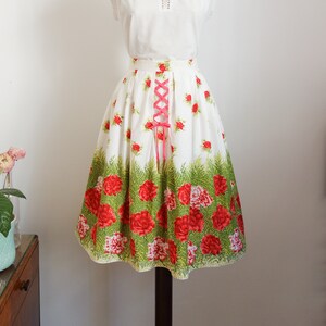 May include: A white skirt with a floral print of red roses and green leaves. The skirt has a pink lace-up detail at the waist.