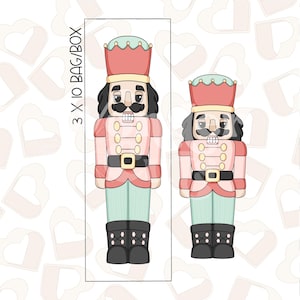 Könnte beinhalten: Zwei Nussknacker-Ausstecher, einer groß und einer klein. Der größere Ausstecher ist rot, weiß und grün mit einem schwarzen Schnurrbart und Bart. Der kleinere Ausstecher ist vom gleichen Design, aber in rosa, weiß und grün. Der Text "3 X 10 BAG/BOX" befindet sich auf der rechten Seite des Bildes.