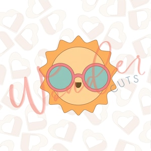 May include: A yellow sun-shaped cookie cutter featuring a smiling face and wearing round pink-rimmed sunglasses with light blue lenses. The design includes a small dark brown mouth and is set against a repeating pattern of heart shapes.