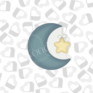 Puede incluir: Cortador de galletas con forma de media luna con una estrella colgando. La luna es de color verde azulado apagado y la estrella es de color amarillo claro. La estrella está unida a la luna con un pequeño lazo.