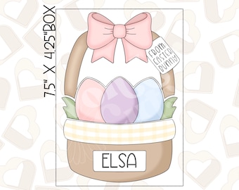 May include: A brown basket cookie cutter with a pink bow, three pastel coloured eggs, and a tag that says "From: Easter Bunny". The basket has a checkered pattern and a name tag that says "Elsa". The cookie cutter is 7.5 inches by 4.25 inches.
