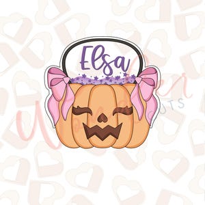 May include: A pumpkin-shaped cookie cutter with a smiling jack-o'-lantern face, pink bows, and the name "Elsa" in purple. The pumpkin is orange with a black outline and a purple star-filled area above it.
