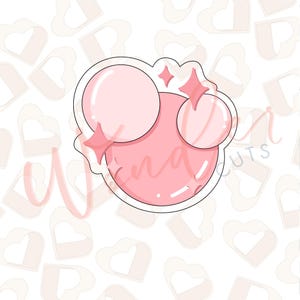 May include: A pink cookie cutter in the shape of a stylized face with three circles and star accents. The cutter has a white outline and is set against a background of hearts and the text "Wena Cuts".