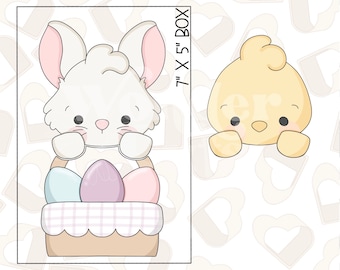 May include: A digital design for a cookie cutter featuring a white bunny holding a basket of pastel Easter eggs. The bunny has pink ears and a pink nose. The basket is brown with a white and pink checkered pattern. The design is 7 inches by 5 inches.