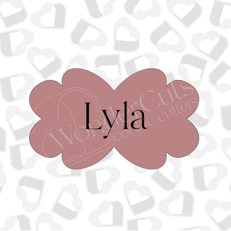May include: A pink cookie cutter in the shape of a bow with the name "Lyla" written in black text. The cookie cutter is on a white background with a pattern of gray hearts.