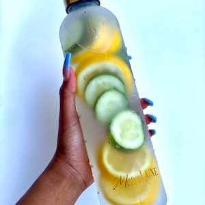 May include: A frosted glass water bottle with a gold lid filled with sliced lemons and cucumbers. The bottle has the text "Meme Luxe Studio" printed on the side.