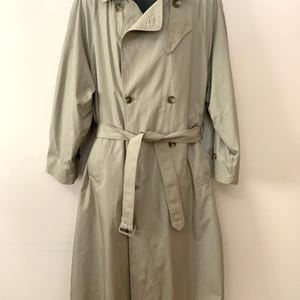 May include: A tan trench coat with a double-breasted design. The coat features a belt at the waist, a notched collar, and button closures. The long sleeves extend to the mid-arm. The coat is displayed on a black mannequin.