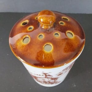May include: A brown ceramic jar with a lid featuring a knob and multiple holes. The jar has a white base with a brown and white floral design.