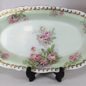 May include: An oval porcelain serving platter featuring a pale mint green background with hand-painted pink rose clusters and green foliage. The scalloped rim is decorated with a delicate gold-painted filigree pattern along the entire edge.