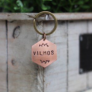 Può includere: Un portachiavi esagonale di colore rame con il nome "VILMOS" inciso su di esso. Il portachiavi è attaccato a un anello portachiavi dorato.