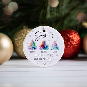 May include: A round white ceramic ornament hanging by a gold string. It features three watercolor-style Christmas trees in rainbow colors, with the text "Sisters", the names "Simone", "Belinda", "Cherie", and the phrase "ARE DIFFERENT TREES FROM THE SAME FOREST" with the year "2025".