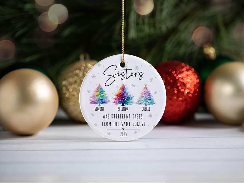 May include: A round white ceramic ornament hanging by a gold string. It features three watercolor-style Christmas trees in rainbow colors, with the text "Sisters", the names "Simone", "Belinda", "Cherie", and the phrase "ARE DIFFERENT TREES FROM THE SAME FOREST" with the year "2025".