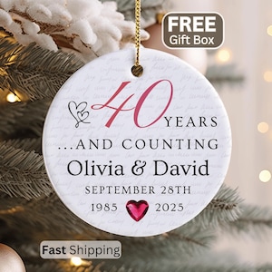 May include: A round white ceramic ornament featuring the text 40 YEARS...AND COUNTING, the names Olivia & David, the date SEPTEMBER 28TH, and the years 1985 and 2025 separated by a small red heart graphic, suspended by a gold string.