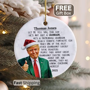 May include: A round white ceramic ornament featuring a portrait of Donald Trump wearing a Santa hat and a green suit with a red tie. The text reads: Thomas Jones, Let me tell you, this guy - he's not just a grandson, he's a tremendous grandson.