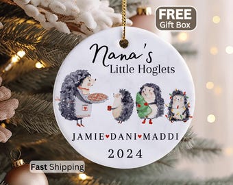 May include: A white ceramic Christmas ornament with a gold string. The ornament features a watercolor illustration of hedgehogs and the text "Nana's Little Hoglets" with names and the year 2024.