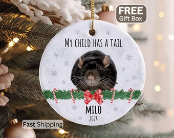 May include: A round white ceramic Christmas ornament featuring a black rat, a green garland with candy canes, and a red bow. The text reads "MY CHILD HAS A TAIL", "MILO", and "2024". The ornament has a marble-like texture and small grey snowflakes.