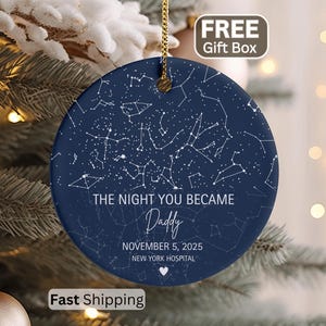 May include: A navy blue ceramic Christmas ornament with a gold string, featuring a constellation design and the text "THE NIGHT YOU BECAME Daddy" with a date and location. The ornament is hanging on a Christmas tree.