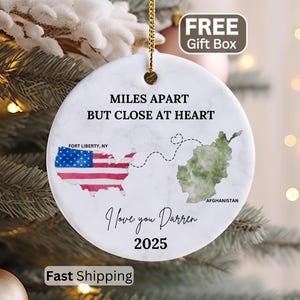 May include: A round white ceramic ornament with a marble texture. It features the text MILES APART BUT CLOSE AT HEART, a map of the USA with a flag pattern, a map of Afghanistan, a dashed line with a heart, I love you Darren, and 2025.