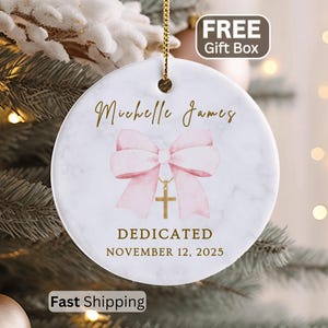 May include: A round white ceramic ornament with a marble texture, featuring a pink bow graphic with a gold cross pendant. The text reads Michelle James, DEDICATED, and NOVEMBER 12, 2025 in gold lettering. It hangs from a gold string.