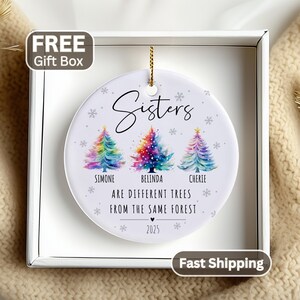 May include: A round white ceramic ornament featuring three colorful watercolor Christmas trees. The text reads Sisters, Simone, Belinda, Cherie, Are different trees from the same forest, and 2025. It is displayed inside a square white gift box.
