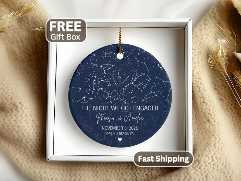 May include: A round, dark blue ceramic ornament featuring a white star constellation map. The text reads: THE NIGHT WE GOT ENGAGED, Mason & Amelia, NOVEMBER 5, 2025, VIRGINIA BEACH, VA, with a small white heart icon at the bottom. It hangs by a gold cord.