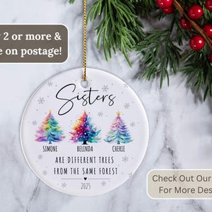 May include: A round white ceramic Christmas ornament featuring three watercolor-style Christmas trees in rainbow colors. The text reads: Sisters are different trees from the same forest, with the names Simone, Belinda, and Cherie, and the year 2025.