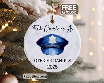 May include: A round white ceramic ornament with a marble texture featuring a blue police hat illustration in the center. The text reads First Christmas As OFFICER DANIELS 2025. The ornament hangs from a gold string against a blurred tree background.