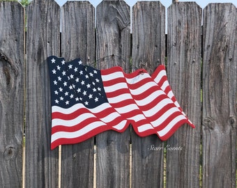 May include: A wooden American flag decoration with red and white stripes and a blue field of white stars. The flag is hanging on a weathered wooden fence. The flag is waving in the wind.
