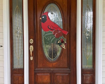 May include: A wooden door with a large red cardinal decoration. The cardinal has a black face and is perched on a branch with red berries and a decorative bow. The door is framed by sidelights with decorative glass.