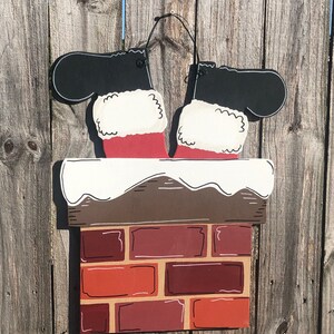 May include: A wooden Christmas decoration depicting Santa's legs and boots in a chimney. The chimney is made of red bricks, and the boots are black with white fur trim. The decoration is hanging on a wooden fence.