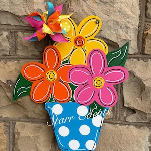 May include: A wooden flower pot decoration with three large, colorful flowers in orange, yellow, and pink. The blue pot has white polka dots and the text "Starr Sefits". A multi-colored pinwheel flower is attached.
