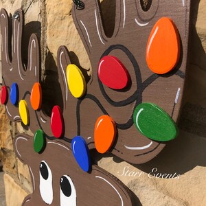 May include: Wooden reindeer head Christmas decoration with colorful lights. The reindeer is painted brown with white accents and a red glitter nose. The lights are painted in various colors, including red, orange, yellow, green, and blue.