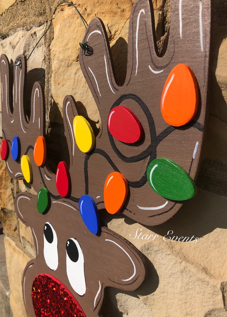 May include: Wooden reindeer head Christmas decoration with colorful lights. The reindeer is painted brown with white accents and a red glitter nose. The lights are painted in various colors, including red, orange, yellow, green, and blue.