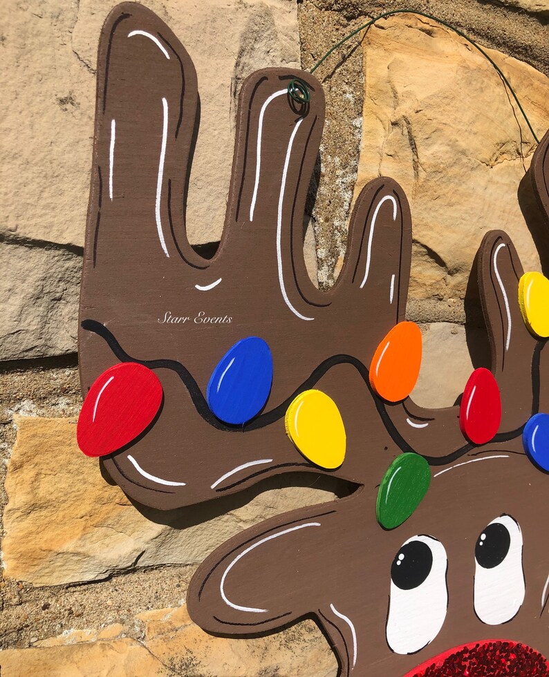 May include: A wooden reindeer Christmas decoration with brown antlers and a smiling face. The antlers are decorated with colorful lights in red, blue, yellow, orange, and green. The reindeer has a red glitter nose.