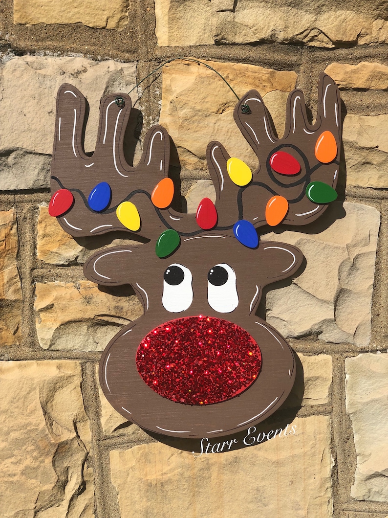 May include: A wooden reindeer Christmas decoration with a brown body, white accents, and a red glitter nose. The antlers are adorned with colorful Christmas lights. The text "Starr Events" is written on the bottom.