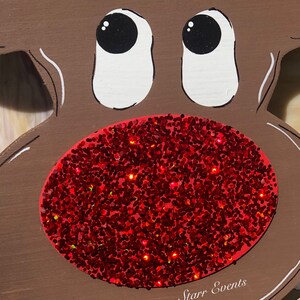 May include: A wooden reindeer face with a large red glitter nose and white-outlined eyes. The reindeer has a brown body with green and blue painted lights on top. The text "Starr Events" is written in white on the bottom.
