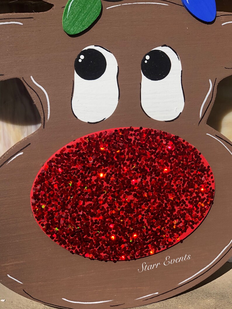 May include: A wooden reindeer face with a large red glitter nose and white-outlined eyes. The reindeer has a brown body with green and blue painted lights on top. The text "Starr Events" is written in white on the bottom.