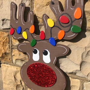 May include: Wooden reindeer Christmas decoration with a brown body, white accents, and a red glitter nose. The antlers are adorned with colorful lights. The decoration is hanging on a stone wall.